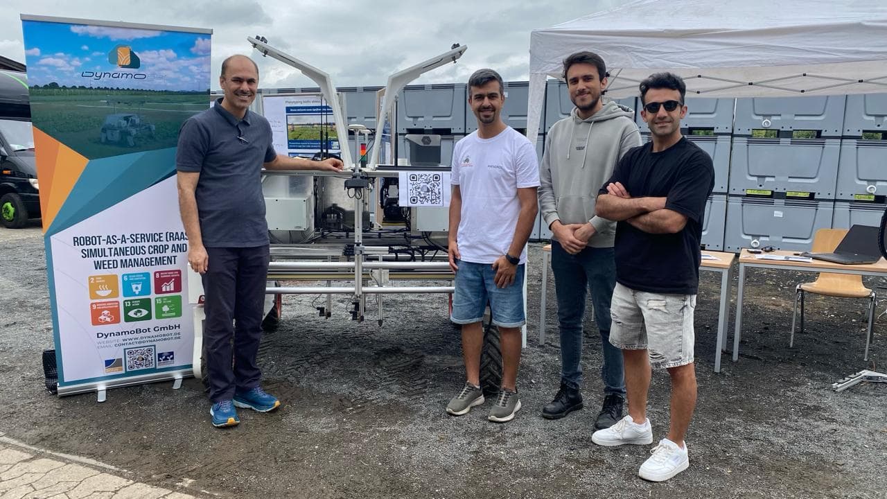 Vier DynamoBot-Teammitglieder vor dem Roboter und einem Banner über Robot-as-a-Service-Unkrautmanagement mit UN-SDG-Symbolen.