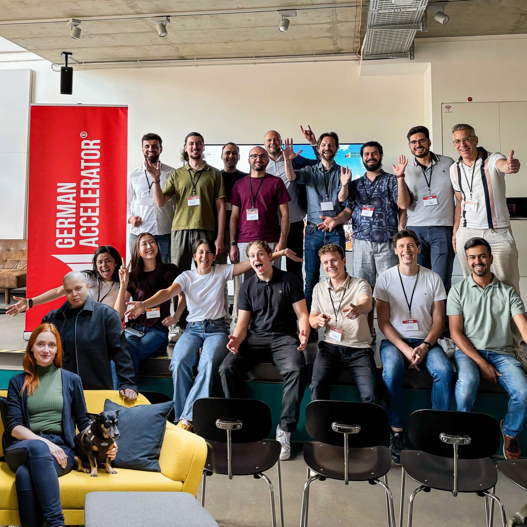 Große Gruppe von Gründerinnen und Gründern unter einem German-Accelerator-Banner in einem Innenraum, einige mit erhobenen Händen.