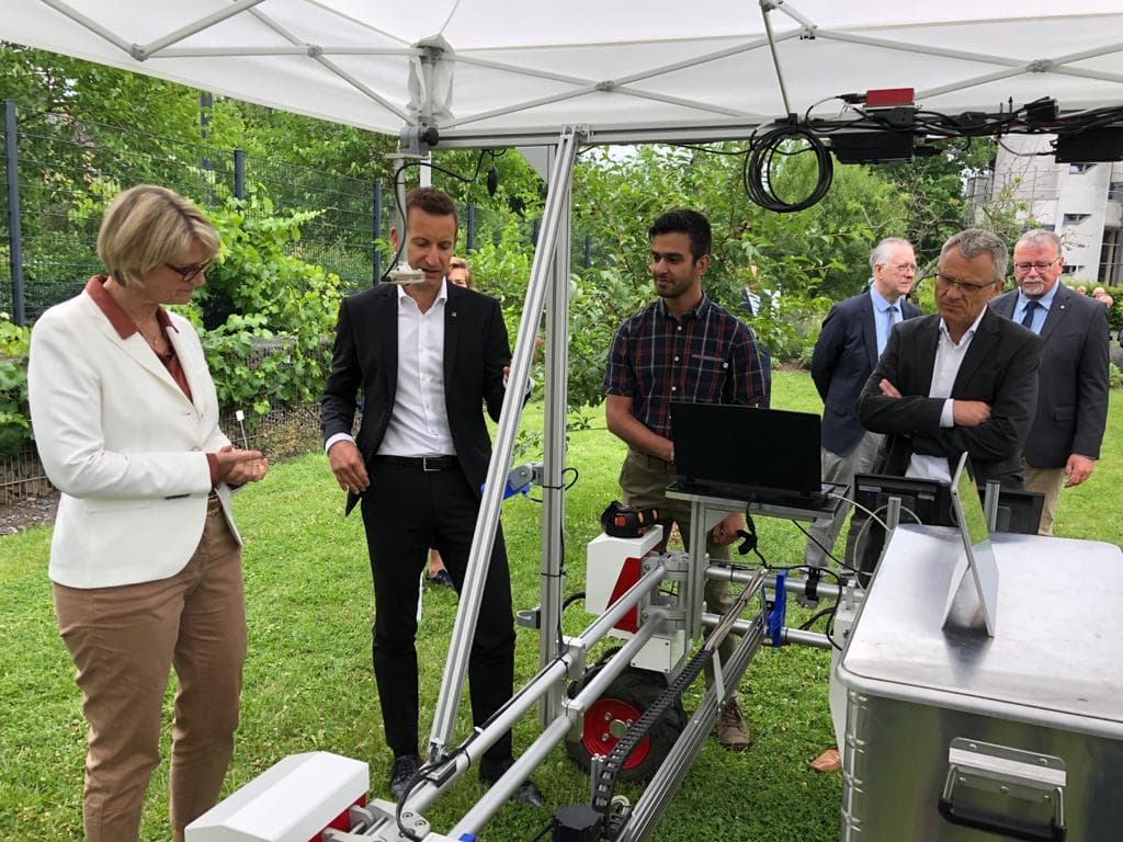 DynamoBot-Roboter mit montiertem Laptop, vorgeführt unter einem Zelt vor einer Gruppe offizieller Gäste in Businesskleidung.