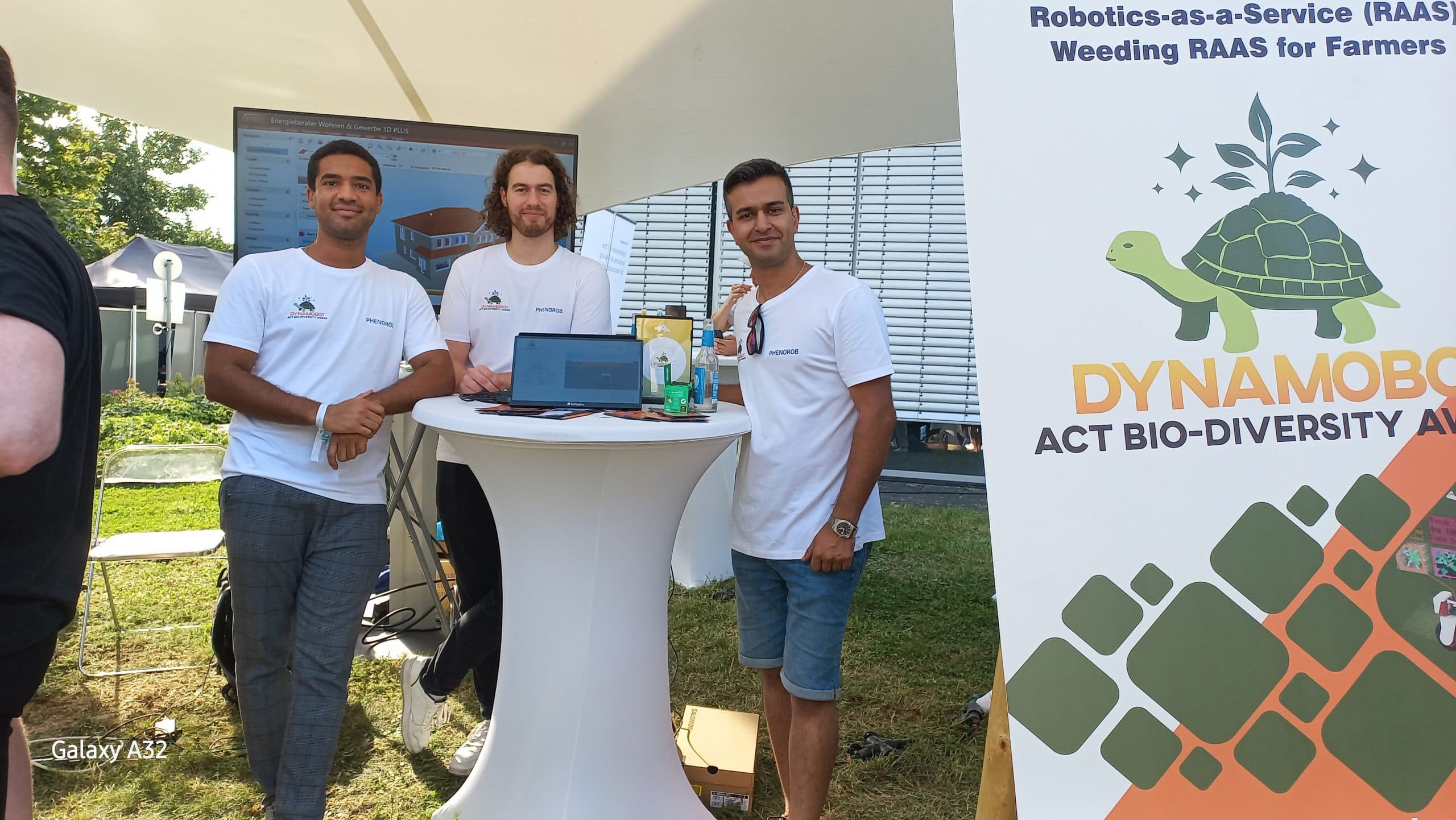 Drei DynamoBot-Teammitglieder an einem Stehtisch neben einem Banner mit der Aufschrift Robotics-as-a-Service-Unkrautmanagement für Landwirte.