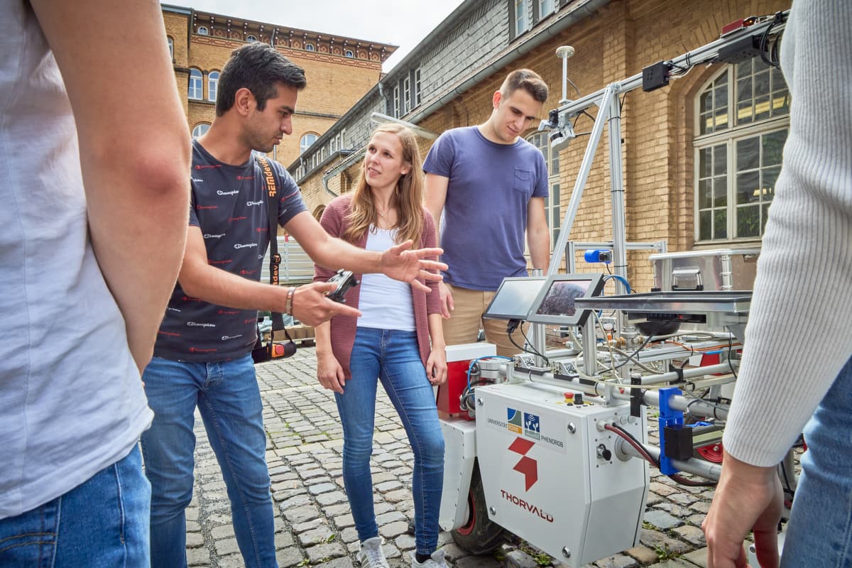 Studierende um den DynamoBot-Roboter in einem gepflasterten Innenhof, während ein Teammitglied ihn erklärt.