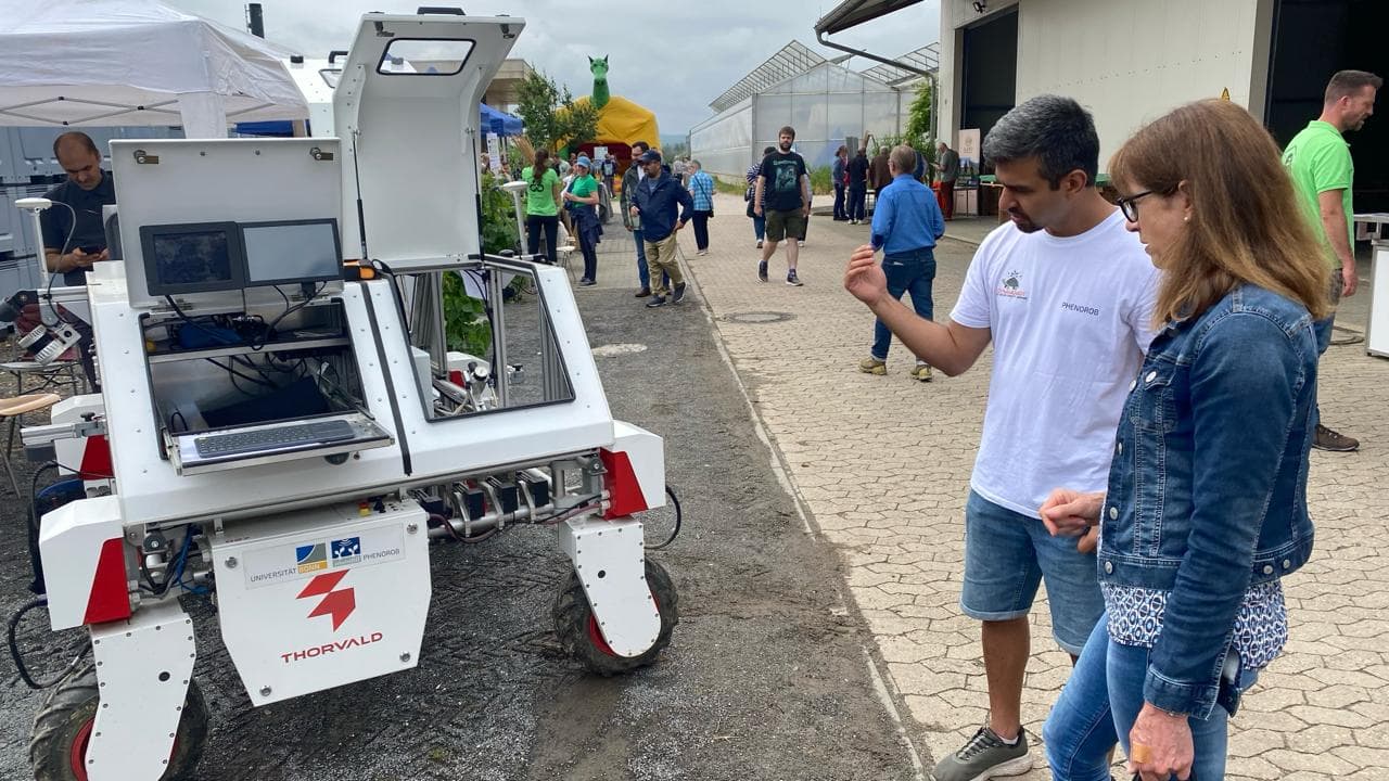 DynamoBot-Jätroboter auf einem gepflasterten Weg auf einem landwirtschaftlichen Feldtag, während zwei Besucher ihn betrachten.