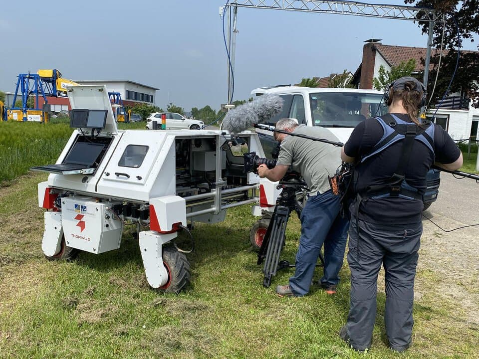 Weißer DynamoBot-Jätroboter auf Gras, während ihn ein Filmteam mit Kamera und Mikrofonangel im Freien aufnimmt.